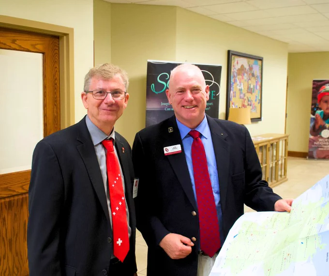 Two individuals in business attire, likely at an event, posing with smiles. They are examining a map placed on a table in front of them.