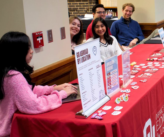 Four individuals seated behind a table, smiling and engaging with visitors. The table is covered with a red cloth and displays brochures, flyers, and promotional buttons.