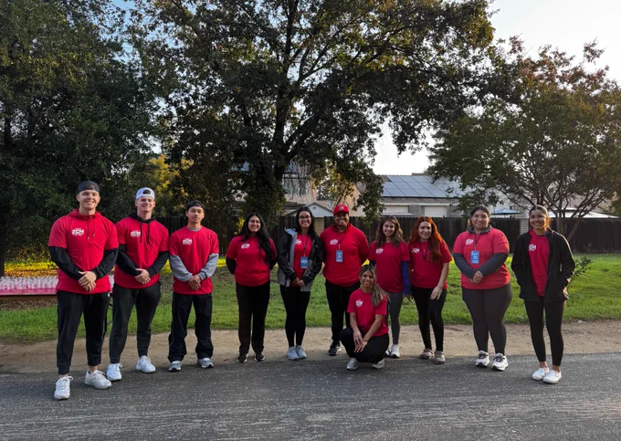Group of people wearing matching red shirts and badges, standing outdoors with trees and a building in the background.