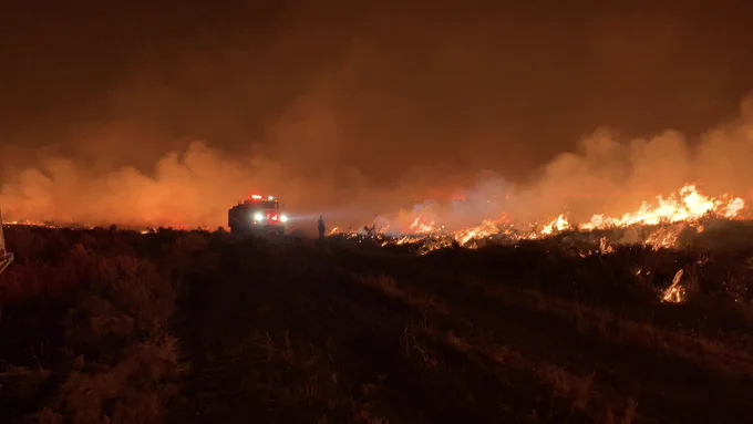 Firefighters battling a large wildfire at night with flames spreading across a dry field under a smoky sky.