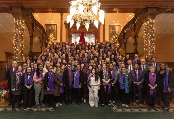 A large group of people, all wearing purple scarves, stand on a grand staircase decorated with festive lights and a red Christmas tree in the background.