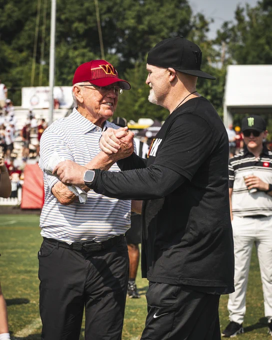Photo of Joe Gibbs and Dan Quinn shaking hands. Gibbs is wearing a burgundy hat and white shirt. Quinn is wearing a black hat and shirt.