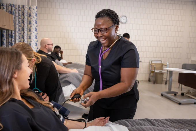 A healthcare professional using a stethoscope to check a patient's heart rate in a clinical setting, both smiling, with other individuals in the background.