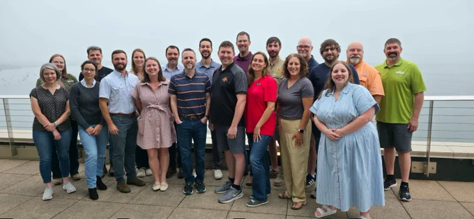 A group of roughly 20 people stands on a balcony overlooking a foggy lake. The group is a mixture of men and women spanning many ages. They are all wearing casual to business casual clothing while standing in two stacked lines.