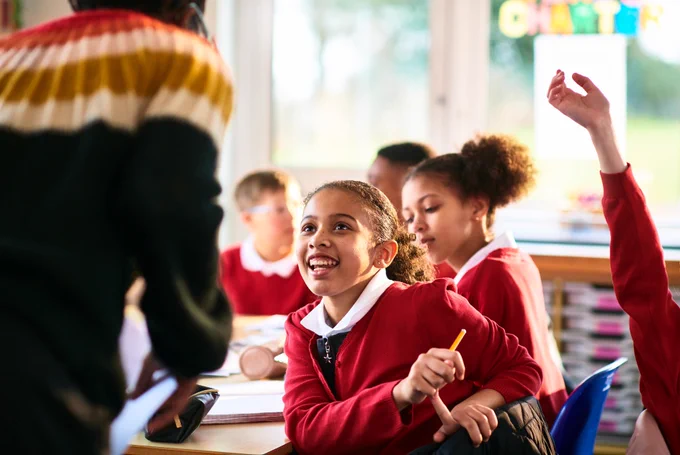 School girl smiles up at her teacher. Other pupils can be seen in the background.