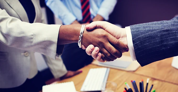 Close-up of a handshake between two people in business attire, symbolizing agreement or partnership. A wooden desk with office supplies is visible in the background."
