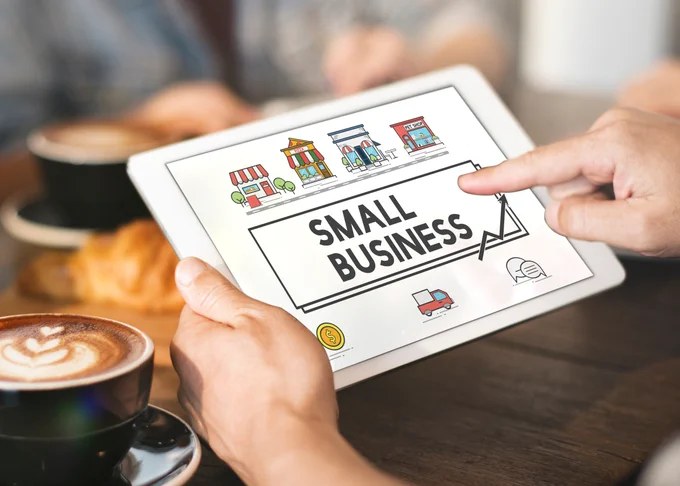 Tablet screen showing the words 'Small Business' with colorful illustrations of storefronts. The image is set in a cafe, suggesting a relaxed working environment for small business owners."