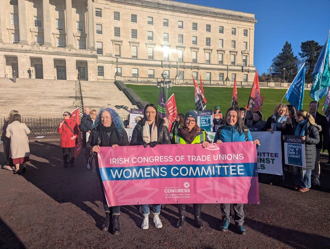 A group of people stand outside Parliament Buildings at Stormont on a bright day, holding a pink banner that reads “Irish Congress of Trade Unions Women’s Committee.” Others behind them hold union flags and signs, including ones from UNISON that say “Where’s Our Safe Leave?”. The crowd is gathered on the steps and forecourt during a union protest.