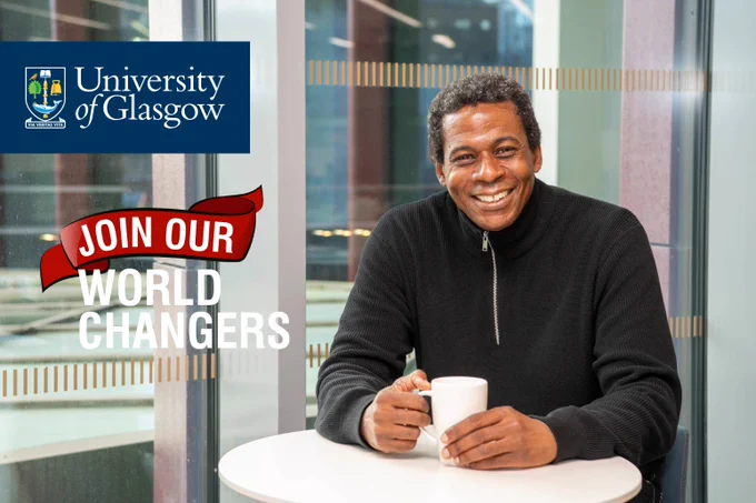 Image shows JMS Scholar Chris Reid in the University of Glasgow’s Advanced Research Centre. He wears a black, quarter-zip jumper and is sitting at a small, round, white table near an upper level window, holding a tall, white mug.