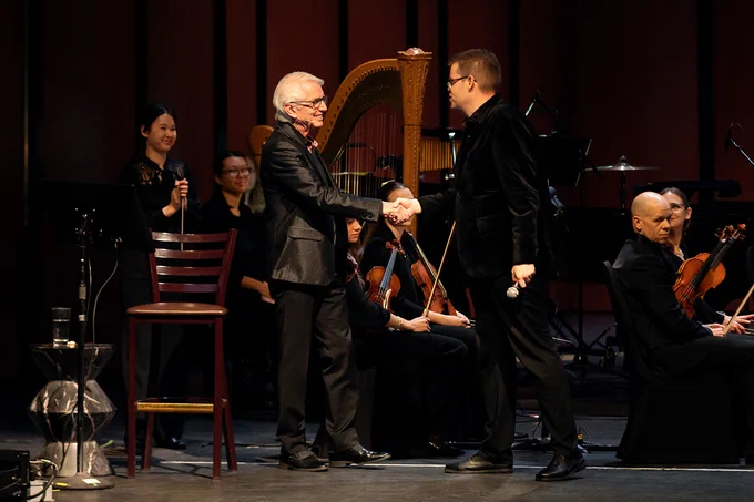 An orchestra performing in front of a large screen displaying a National Geographic image of a whale breaching the ocean surface, titled "Secrets of the Whales."
