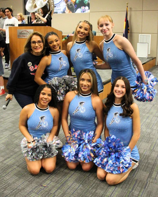 Group of cheerleaders in blue and silver uniforms with pom-poms, posing together indoors.