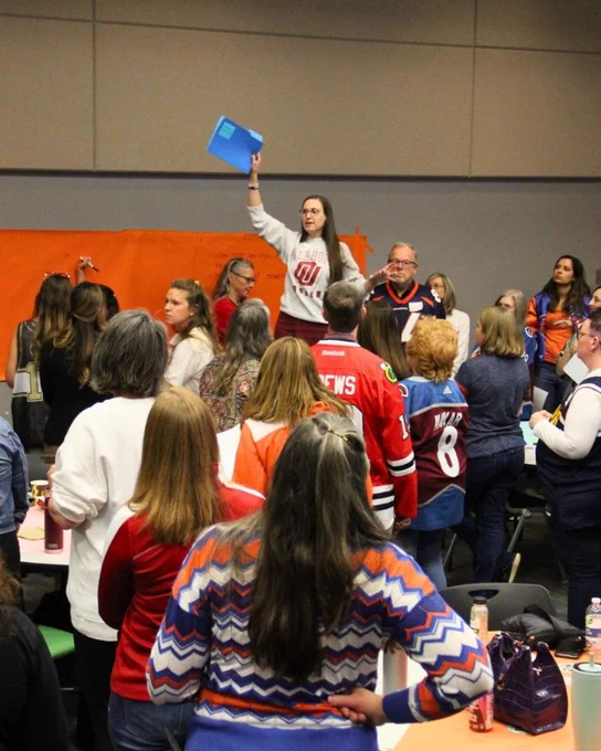 A person stands raising a blue book above their head in a crowded room where attendees wear various colorful outfits, including sports jerseys.