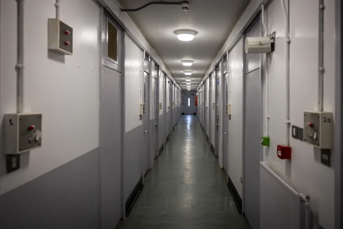 View down a prison corridor, with white walls and grey cell doors on both sides.