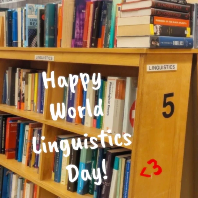 Happy World Linguistics Day! <3 in front of a wooden bookshelf with the label "linguistics" 