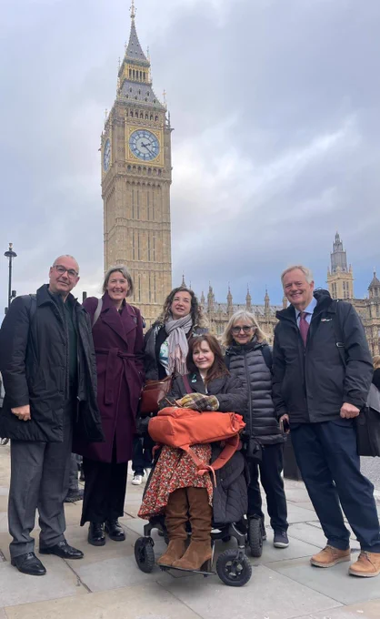 A group of six people stand and smile in front of the Houses of Parliament in London, with the Elizabeth Tower (Big Ben) visible behind them. One person is seated in a wheelchair at the centre. They are dressed in winter clothing and appear to have just come from a meeting or event. The sky is overcast.