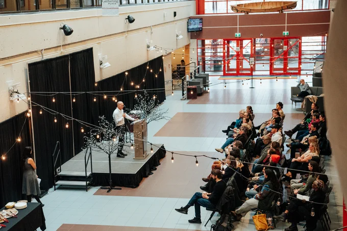 Speaker addressing an audience at a podium in an indoor atrium decorated with string lights.