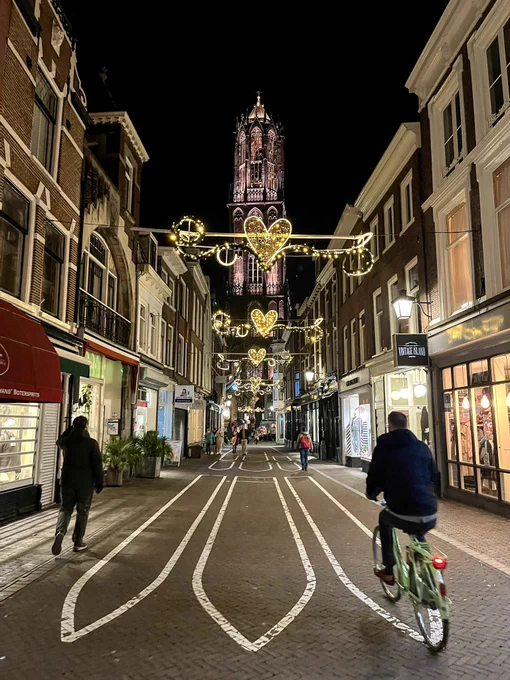 A cyclist passes through an empty street in the medieval centre of Utrecht after dark, with Christmas lights overhead and the Dom Tower in the background.