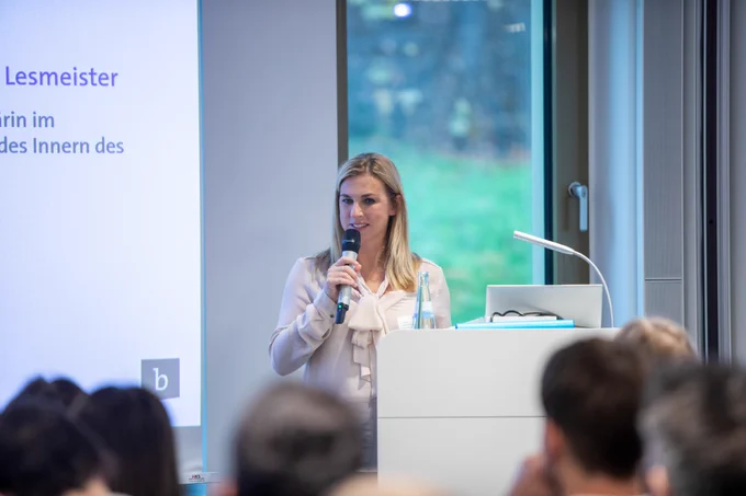 Daniela Lesmeister, StS bei Innenministerium NRW spricht bei der Jahrestagung der Sicherheitskooperation Cybercrime 2025 am Rednerpult mit Mikrofon, im Hintergrund Präsentationsfolie und Fenster mit Ausblick ins Grüne. Im Vordergrund sind unscharf die Köpfe mehrerer Zuhörender zu erkennen.