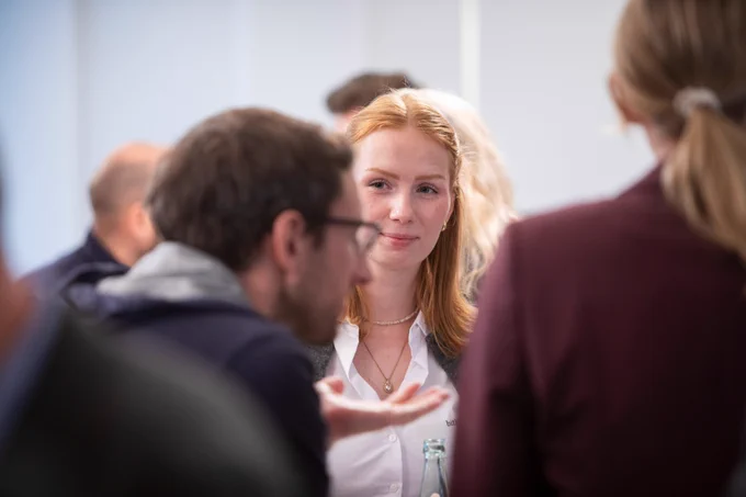Eine junge Frau mit schulterlangem rotem Haar steht inmitten einer Gruppe von Menschen in einem Gespräch auf der der Jahrestagung der Sicherheitskooperation Cybercrime 2025. Die Frau schaut aufmerksam auf einen Mann im Vordergrund, der sich im Gespräch befindet. Um sie herum stehen mehrere Personen, deren Gesichter teilweise unscharf sind.