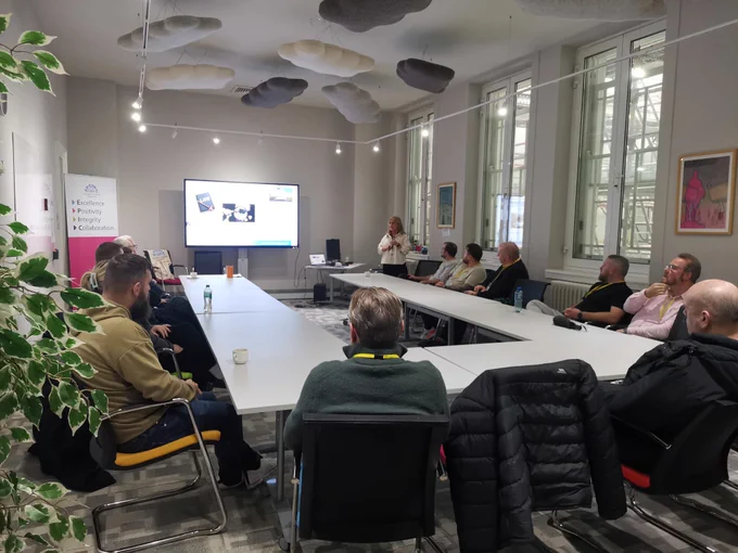 The photo shows a group of 15 people sitting at tables in a room. The participants are looking at a large screen, with a presenter standing at the front talking.