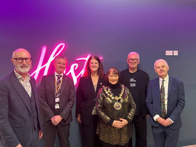 A group of six people standing in front of a dark blue wall with a bright pink neon sign that reads “Host.” The individuals are dressed in formal and semi-formal attire, including suits and a ceremonial chain worn by one person in the center. The setting appears to be an indoor event space with modern decor.