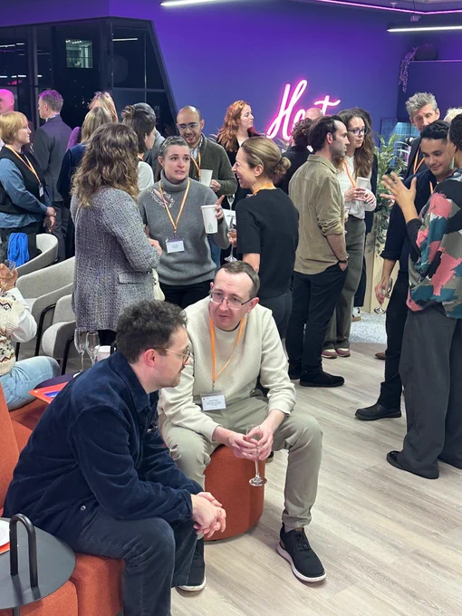 A lively networking event with numerous attendees gathered in a modern indoor space. People are standing and conversing in small groups, some holding drinks and wearing name badges with orange lanyards. In the foreground, two individuals are seated on orange cushioned furniture, engaged in conversation. A pink neon sign reading “Host” is visible on the wall in the background.