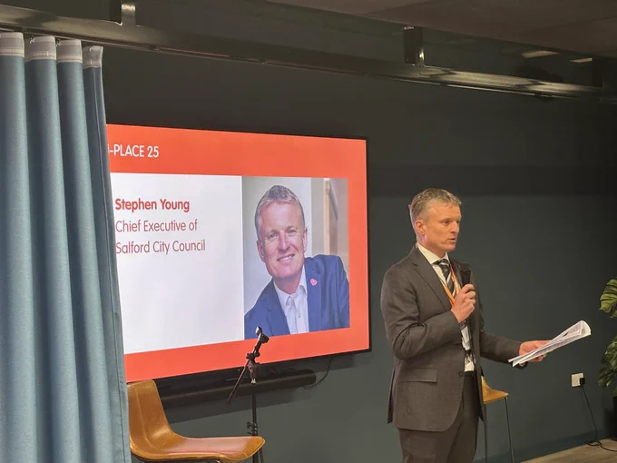 Stephen Young, CEO Salford Ciity Council, speaking into a microphone while holding papers, standing next to a large screen. The screen displays a presentation slide with the text “Stephen Young, Chief Executive of Salford City Council” alongside a portrait photo. The setting includes a modern interior with a curtain, chairs, and a plant visible in the background.