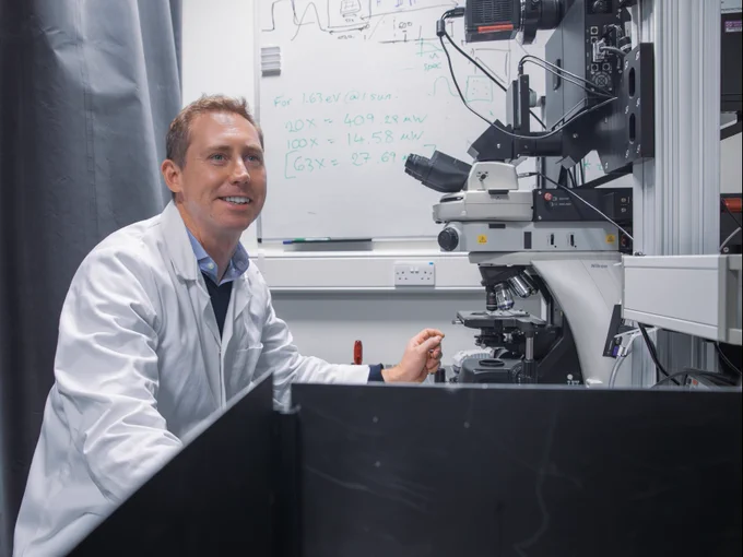 Professor Sam Stranks in a lab coat smiles while working with a microscope, surrounded by advanced laboratory equipment. Whiteboards with mathematical equations are visible in the background.