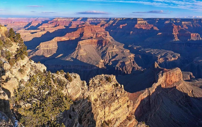 View down past a limestone ridgeline dotted with trees. In the distance colorful peaks and cliffs rise from the floor of a mile deep canyon