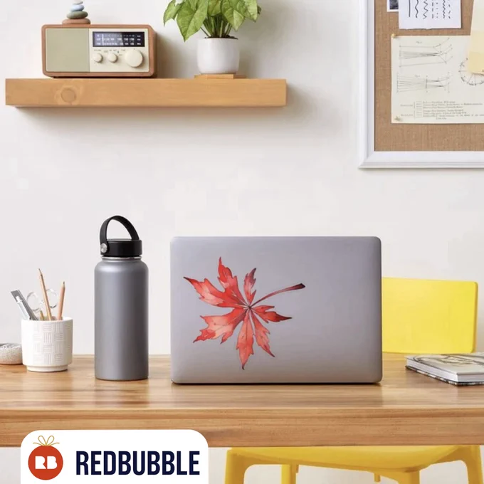 A silver laptop on a wooden desk shows a red watercolor maple leaf sticker in the center of the lid. Next to it stands a gray water bottle and a cup filled with pencils. A yellow chair and soft neutral decor create a calm workspace atmosphere. The focus is on the vibrant red leaf sticker as a decorative accessory for tech items.