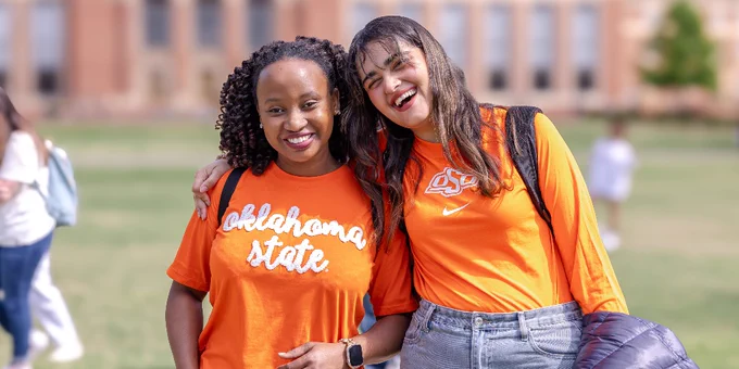 Two individuals in Oklahoma State University attire, smiling happily with linked arms, standing outdoors.