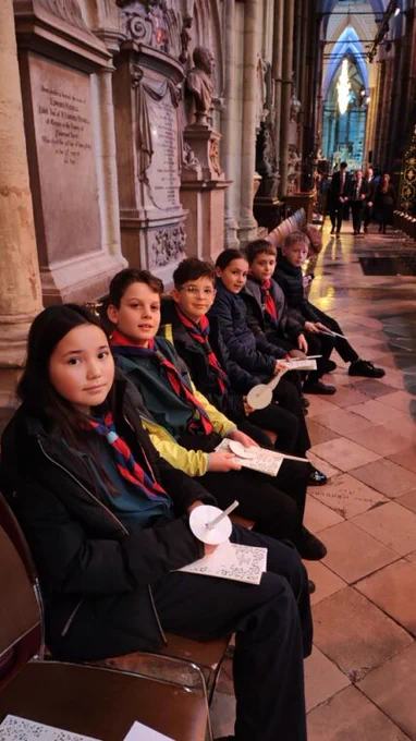 Scouts in uniform sitting on a bench inside of Westminster Abbey, holding candles. 