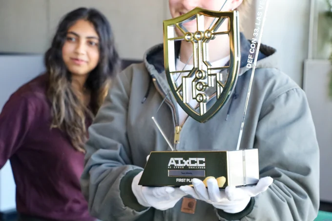 A man wearing white gloves holds a trophy. It says AIxCC First Place Team Atlanta. Def Con 33, Las Vegas. A woman stands behind him looking out of the frame.