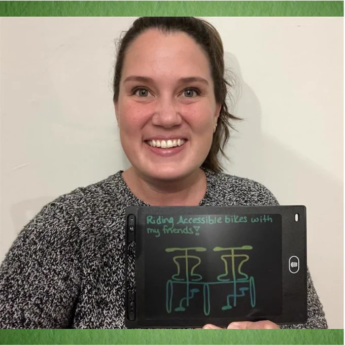 A person is smiling and holding a sketch tablet showing a drawing of an adapted bicycle with the phrase “Riding accessible bikes with my friends!”