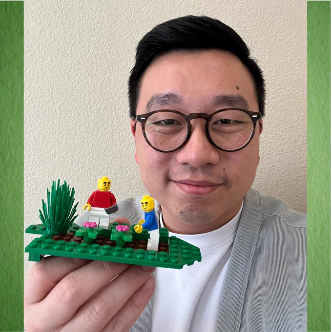 A person smiles while holding a small Lego construction of 2 people gardening. 