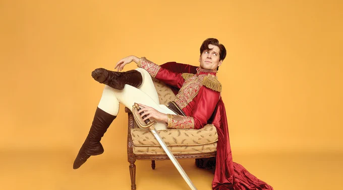 Man dressed in an ornate red and gold royal costume reclining on a vintage armchair against a solid orange background.