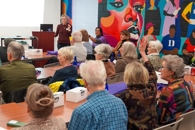 Photographed from the back, the image shows multiple people sitting at tables indoors. At the front of the room, a person stands near a podium, speaking. A painted mural is on the wall behind the speaker.