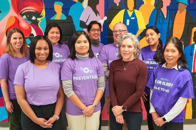 A group of nine people posing for a photo. They are standing in front of a mural. Most of the people in the photo are wearing purple shirts.