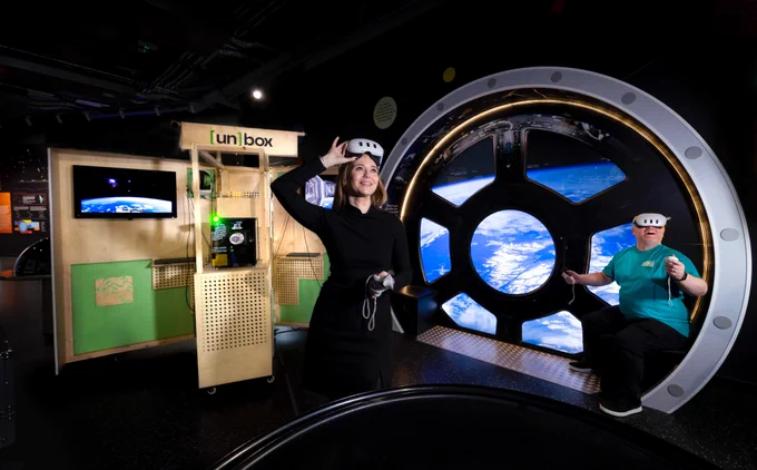 Two people use virtual-reality headsets in a space-themed exhibition. A woman in a black outfit stands smiling while lifting a VR headset from her eyes, holding a controller in her other hand. Behind her is a wooden display unit with a screen showing an image of Earth from space. To the right, a man wearing a teal shirt sits inside a large circular window structure designed to look like a spacecraft cupola, with a projected view of Earth below. 