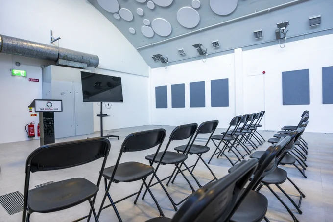A large room with rows of black folding chairs facing a large TV screen. Acoustic panels adorn the walls and ceiling. There is a lectern with The Digital Hub's logo on the front. The walls are painted white and the floor is grey.