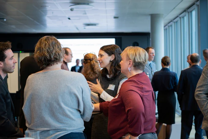 Mehrere Personen stehen in einem modernen Konferenzraum zusammen und unterhalten sich angeregt. Im Vordergrund befinden sich vier Frauen und ein Mann, die in Kleingruppen kommunizieren. Eine Frau mit langen dunklen Haaren, grauem Kleid und weißer Bluse spricht mit einer Frau im bordeauxroten Oberteil. Der Raum ist hell, mit großen Fenstern auf der rechten Seite. Im Hintergrund sind weitere Personen im Gespräch vertieft.