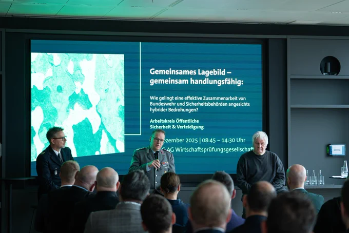 Die drei Speaker Oberst i.G. Andreas Schreiber, DPB Roman Seifert, Leiter der Landespolizeidirektion Berlin und Albrecht Broemme sitzen auf einer Bühne vor einem großen Bildschirm, der eine Präsentation zeigt. Oberst i.G. Andreas Schreiber in der Mitte trägt eine Bundeswehr-Uniform und spricht in ein Mikrofon. Das Publikum ist von hinten zu sehen. Der Bildschirm zeigt den Titel: „Gemeinsames Lagebild – gemeinsam handlungsfähig: Wie gelingt eine effektive Zusammenarbeit von Bundeswehr und Sicherheitsbehörden angesichts hybrider Bedrohungen?“. 