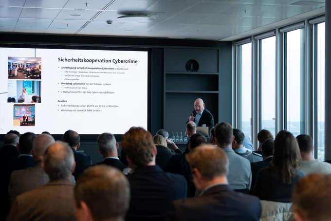 Uwe Sanders, Vorsitzender des Bitkom-AK Öffentliche Sicherheit spricht mit einem Mikrofon an einem Rednerpult. Er steht neben einem großen Bildschirm, auf dem der Titel „Sicherheitskooperation Cybercrime“ und verschiedene Punkte zu Veranstaltungen und Ausblicken aufgelistet sind. Das Publikum sitzt in Reihen und schaut nach vorne. Rechts ist eine große Fensterfront mit Blick auf eine Stadt.