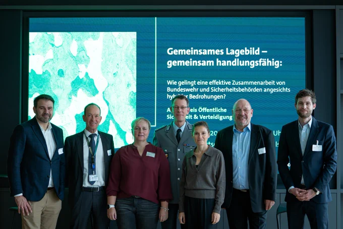 Sieben Personen stehen nebeneinander vor einem großen Bildschirm und posieren für ein Gruppenfoto. Drei Männer in Anzügen, eine Frau in bordeauxroter Bluse, eine Frau in grauem Pullover sowie zwei Männer in Bundeswehr-Uniform. Im Hintergrund ist der Titel der Veranstaltung „Gemeinsames Lagebild – gemeinsam handlungsfähig“ zu lesen.