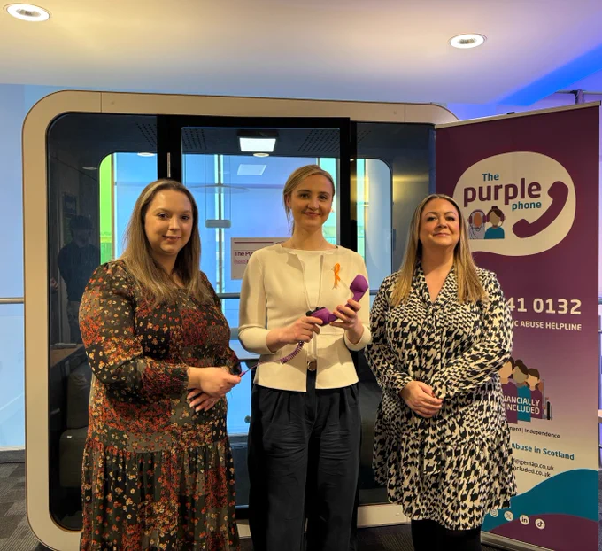 Housing Secretary Màiri McAllan (centre) holding a purple phone with Project Manager at Financially Included Amber Cully  (right) and Councillor Katie Pragnell, Chair of East Renfrewshire Integration Joint Board (left).