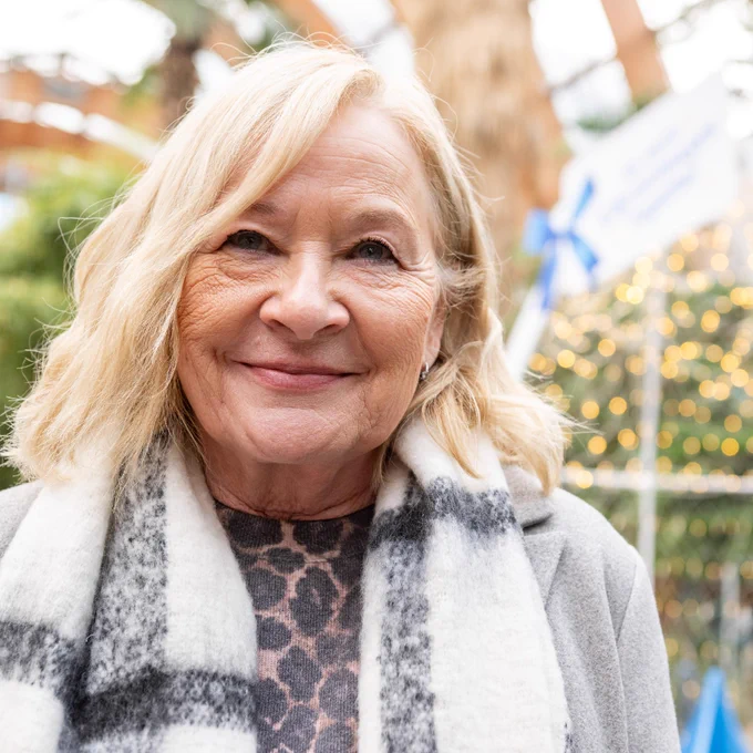 A close-up photo of Lesley wearing a scarf and grey coat, standing in Sheffield Winter Gardens with Christmas lights in the background.