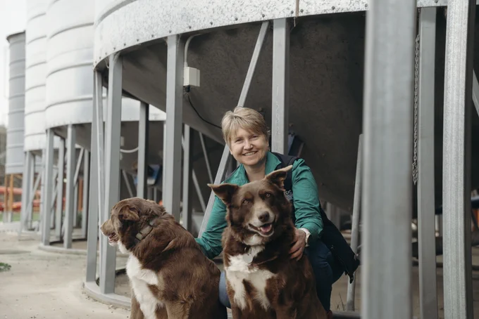 A person is smiling while seated outside next to two dogs at a farm with silos in the background.