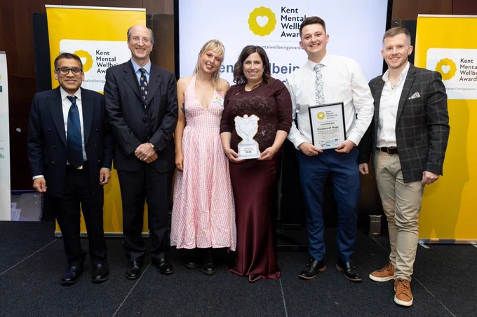 Six people are stood in a row in front of a Kent Mental Wellbeing Awards banner. They are smiling and two are holing a wooden trophy and an award-winners certificate.