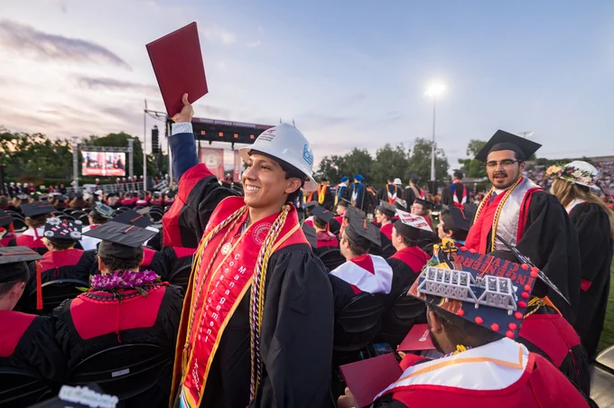 Graduates celebrating at an outdoor ceremony, some wearing decorated mortarboards, with one person joyfully holding up a diploma.