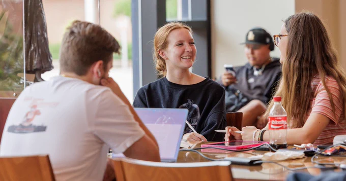 Three students sitting at a table, engaging in a friendly conversation in a campus setting. 